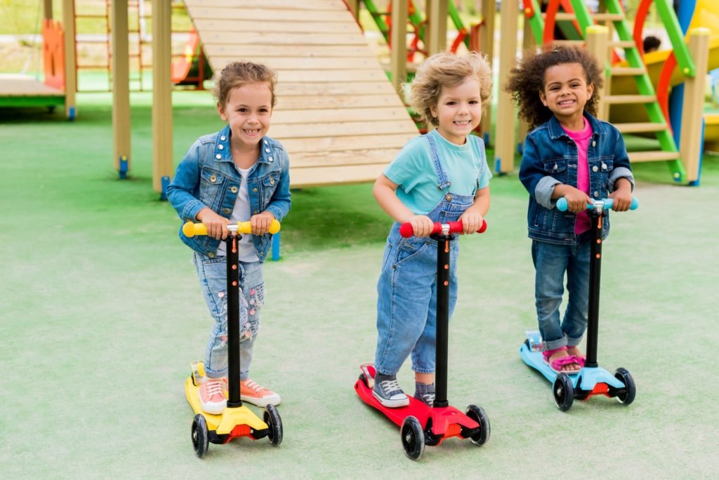 three multicultural adorable little children riding on kick scooters at playground